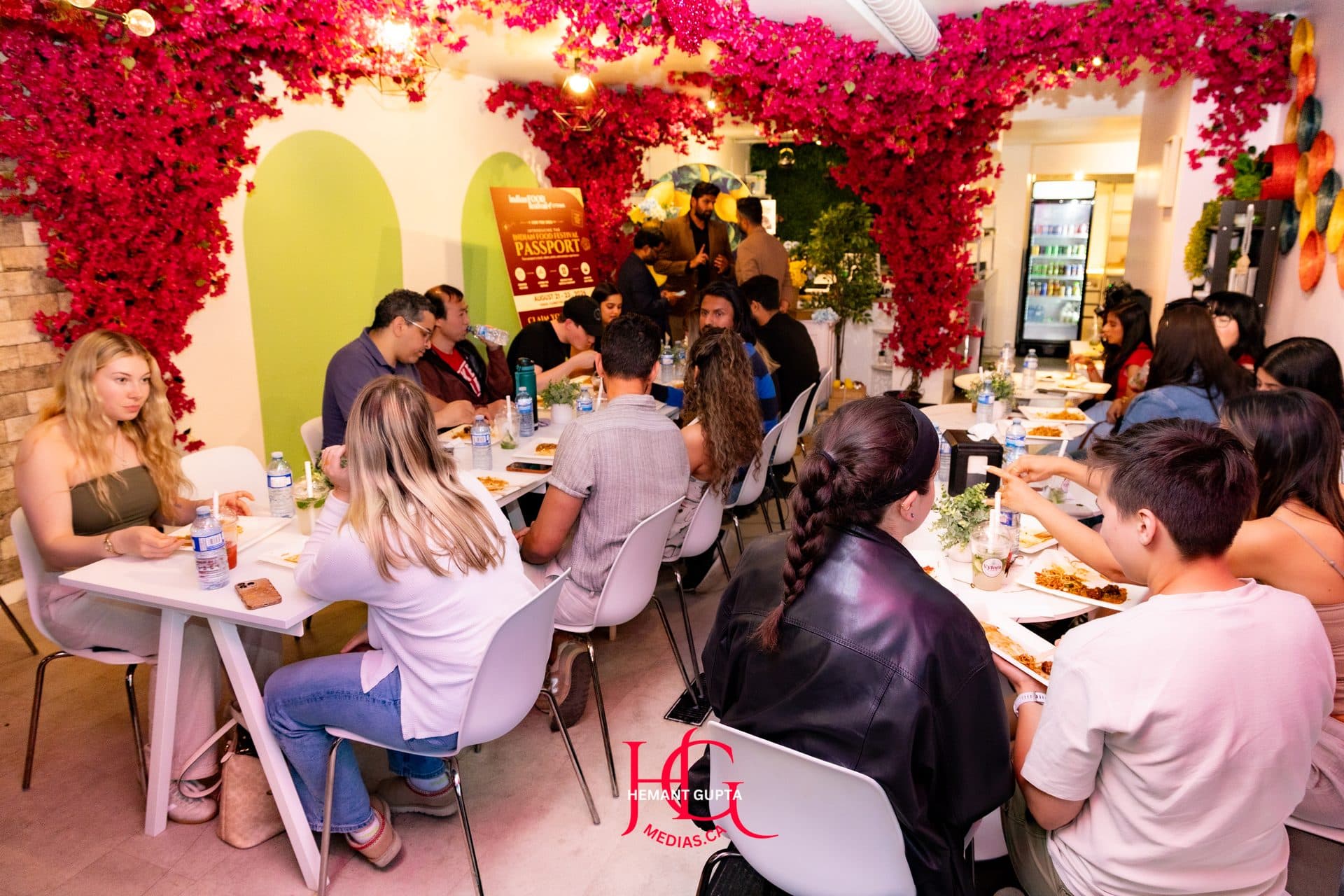 Guests seated and dining together beneath a floral archway, with the Festival Passport poster in the background