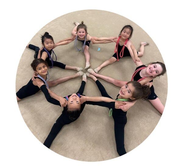 Seven young gymnasts in black leotards posing in a star formation, each holding a rhythmic gymnastics ribbon